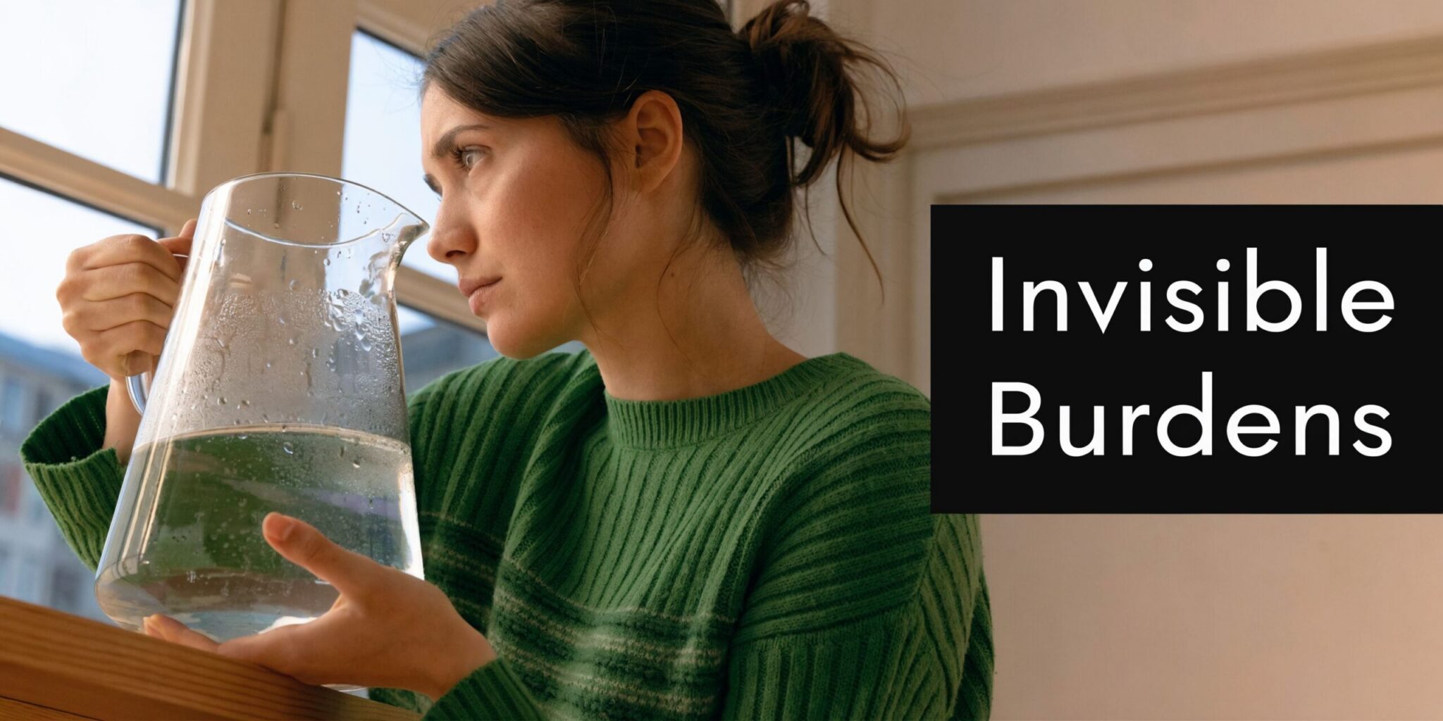 A contemplative woman in a green sweater holding a glass pitcher of water near a window.