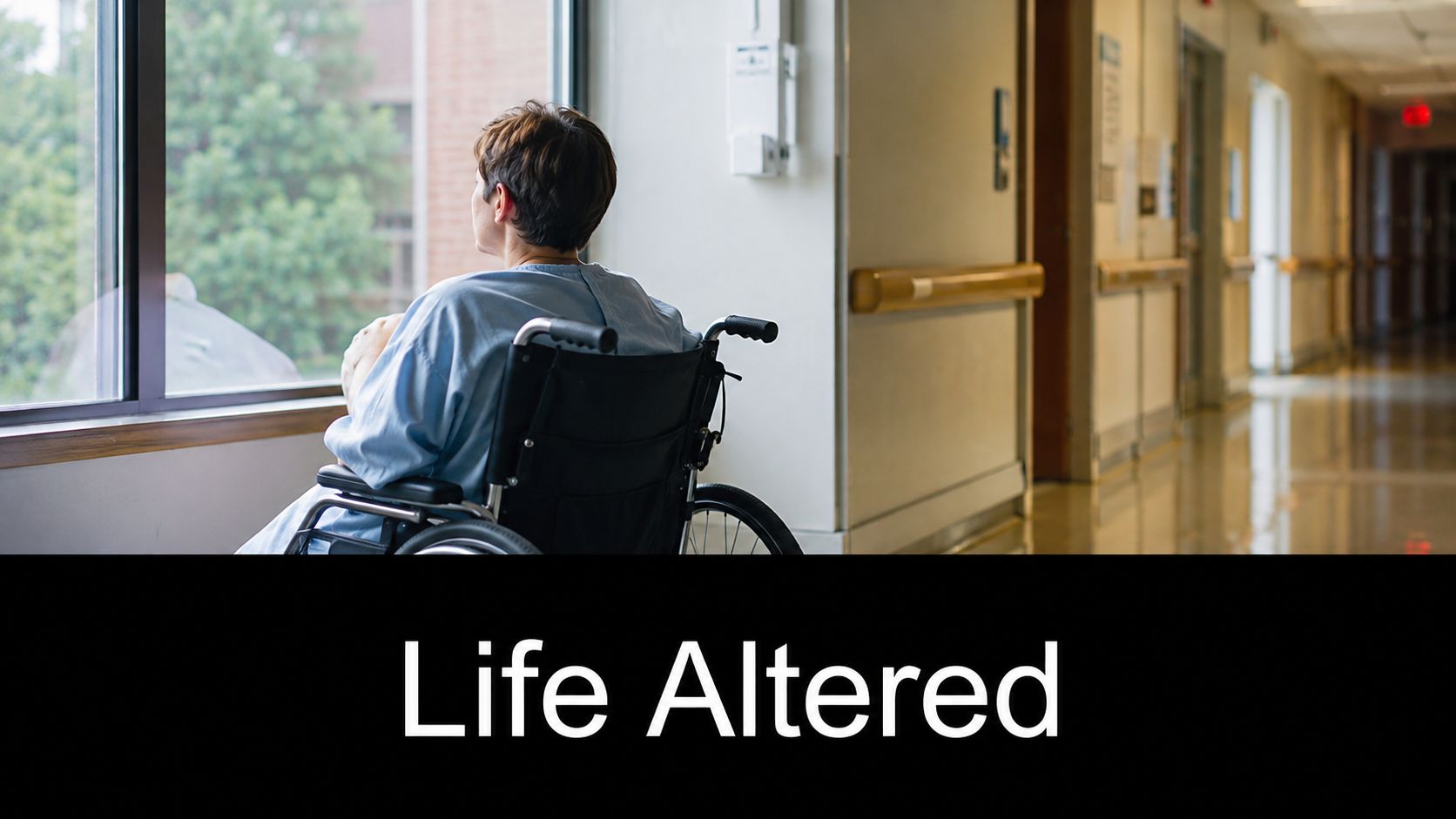 A patient in a hospital gown sitting in a wheelchair looking out a window at a hospital hallway.